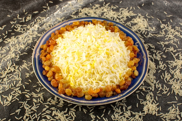 A top view tasty pilaf with oil and dried raisins inside plate with raw rice on the dark surface