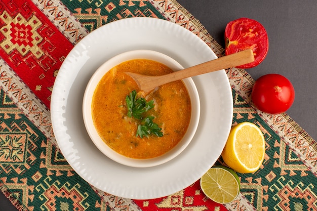 Top view of tasty vegetable soup inside plate with tomatoes and lemon on dark surface