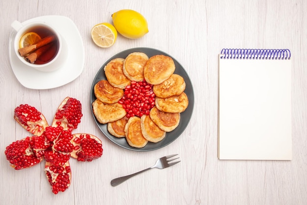 Free photo top view tea and pancakes a cup of tea with cinnamon and lemon peeled pomegranate lemon next to the white notebook plate of seeds of red pomegranate and pancakes and fork on the white background