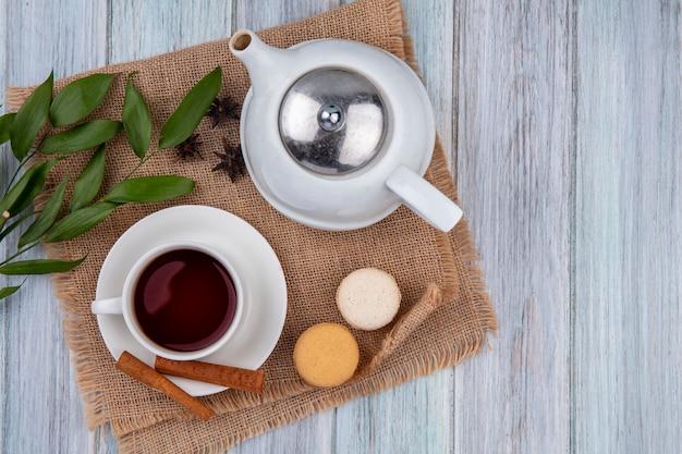 Top view teapot with a cup of tea cinnamon and macaroons on a beige napkin on a gray table