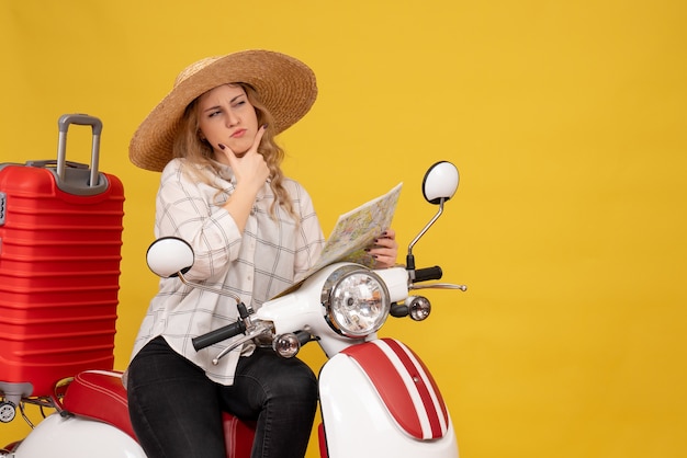 Top view of thinking young woman wearing hat and sitting on motorcycle and holding map concentrated on something on yellow 