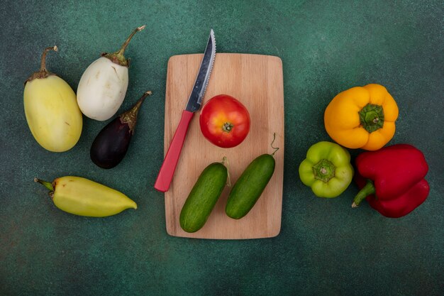 Top view tomato with cucumbers on a cutting board with a knife and colored bell peppers  eggplants on a green background