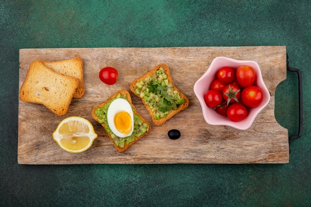 Top view of tomatoes on a pink bowl on a wooden kitchen board with a toasted slices of bread with avocado pulp and egg on green surface