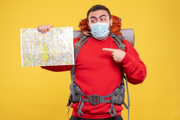 Top view of a traveller guy wearing medical mask with backpack pointing map on yellow background