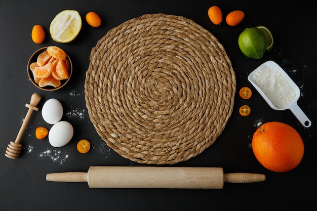 Top view of trivet with egg lemon lime orange tangerine kumquat flour with honey dipper and rolling pin on black background