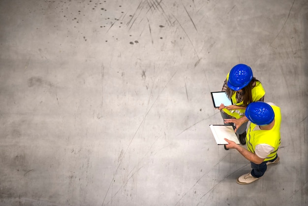 Free photo top view of two industrial workers wearing hardhats and reflective jackets holding tablet and checklist on gray concrete floor