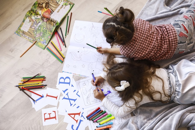 Top view of two tiny girls which are drawing in the coloring book laying on the floor on the blanket