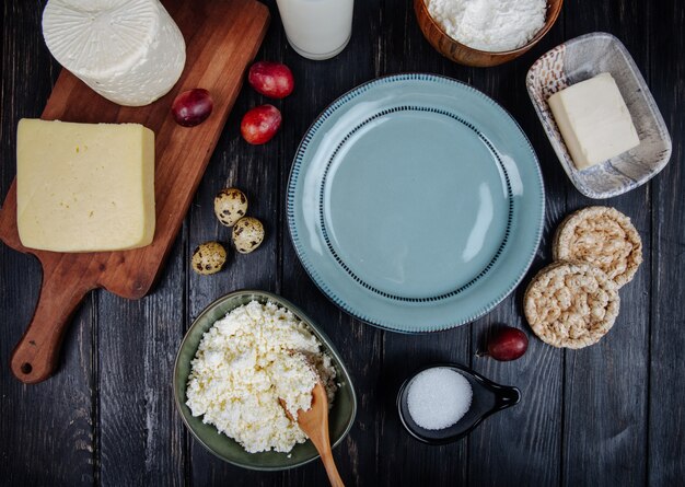 Top view of various kind of cheese with cottage cheese in a bowl, quail eggs, fresh sweet grapes and an empty plate on dark wooden table