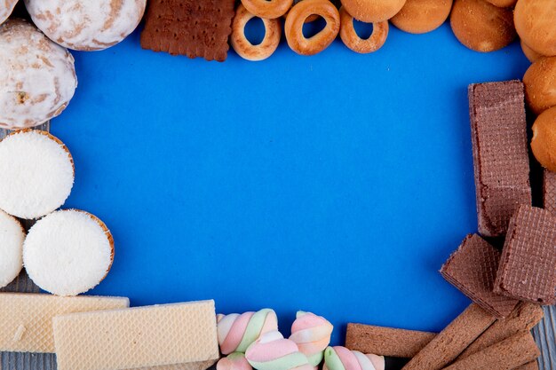 Top view of various types of sweet cookies marshmallows and bread rings arranged in a shape of frame on blue background with copy space