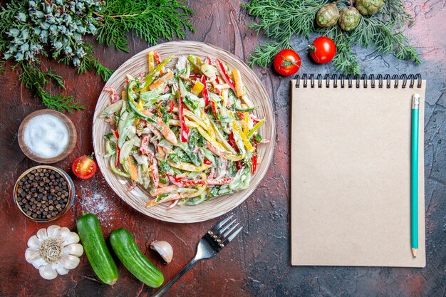 Top view vegetable salad on plate fork tomatoes pine branches cucumbers garlic pencil on notepad on dark red table