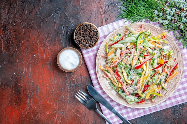 Free Photo top view vegetable salad on plate on tablecloth fork and knife salt and black pepper on dark red table free space