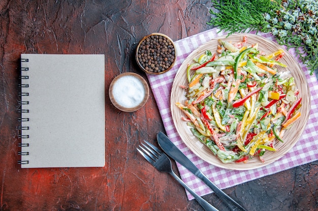 Free photo top view vegetable salad on plate on tablecloth fork and knife salt and black pepper a notebook on dark red table