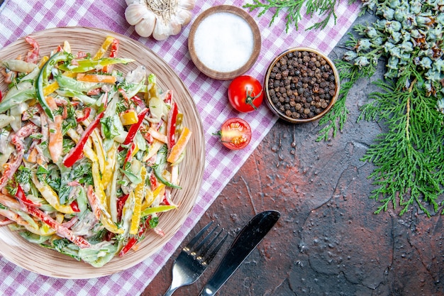 Top view vegetable salad on plate on tablecloth fork and knife salt and black pepper pine branch on dark red table