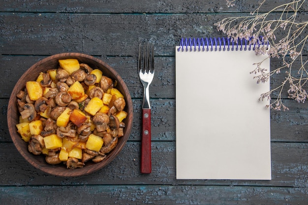 Free photo top view vegetable with mushrooms potatoes and mushrooms in the bowl next to the fork notebook and branches on the dark table