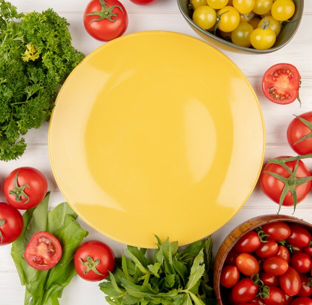 Top view of vegetables as coriander tomato spinach green mint leaves with empty plate on center on wooden surface