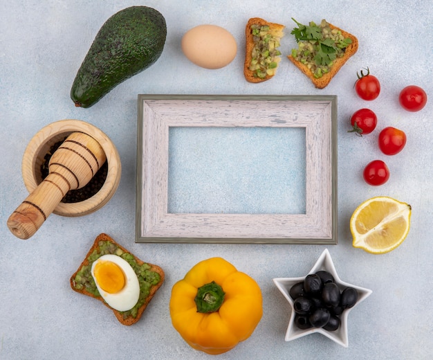 Free photo top view of vegetables such as avocado lemon black olives yellow bell pepper tomatoes and a bread slice with avocado pulp on white surface