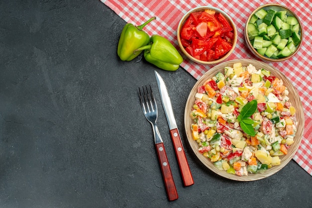 Top view of veggie salad in bowl with red napkin under it and cutleries with vegetables on side and place for text on dark grey background