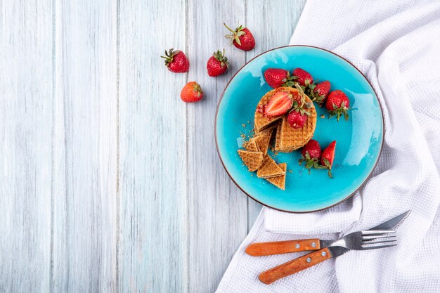 Top view of waffle biscuits and strawberries in plate with fork and knife on cloth and wooden surface