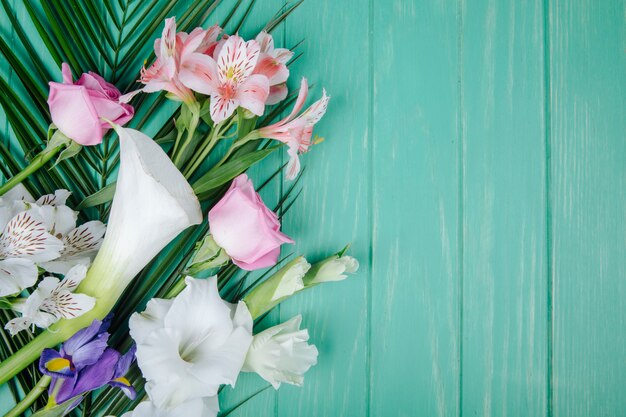 Top view of white color calla lilies and gladiolus with dark purple iris and pink roses and alstroemeria flowers on palm leaf on green wooden background with copy space