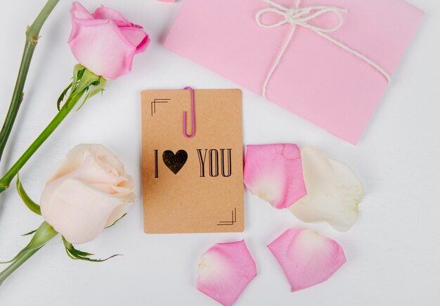 Top view of white and pink color roses with envelope tied with a rope and small postcard with a paper clip and rose flower petals on white background