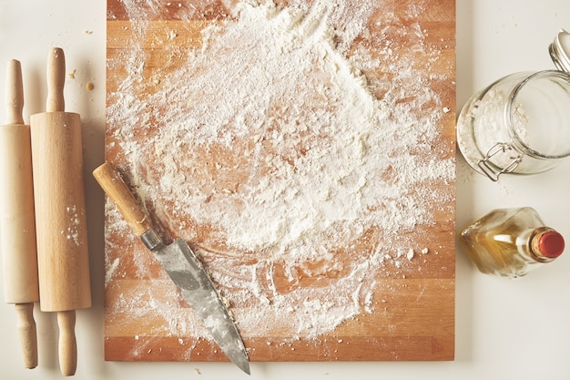 Top view on white table with isolated wooden board with knife, two rolling pins, bottle olive oil, transparent jar with flour. Presentation cooking process