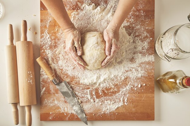 Top view on white table with isolated wooden board with knife, two rolling pins, bottle olive oil, transparent jar with flour Woman hands hold prepared dough for pasta or dumplings