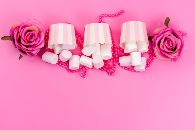 A top view white tasty marshmallows inside paper packages with pink roses on pink desk, biscuit meringue sweet