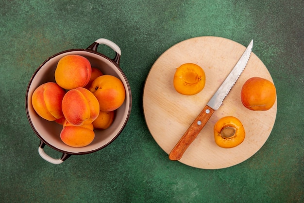 Free photo top view of whole apricots in bowl and half cut one with knife on cutting board on green background