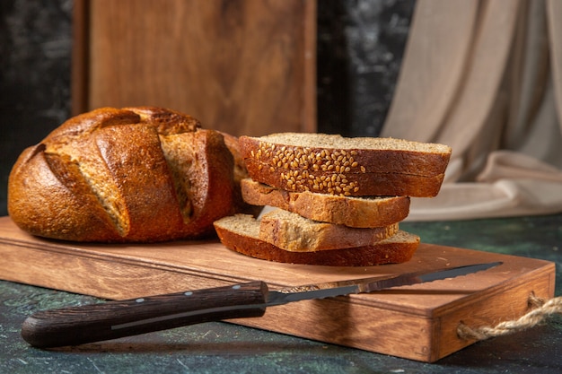 Top view of whole and cut black bread on brown wooden cutting board on dark surface