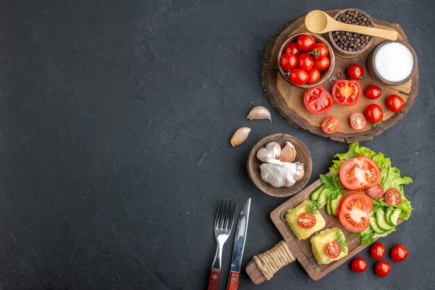 Top view of whole cut fresh vegetables and spices on wooden board white towel cutlery set cheese on black surface