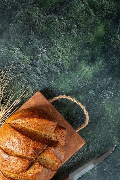 Top view of whole fresh black bread on brown wooden cutting board knife on dark colors surface