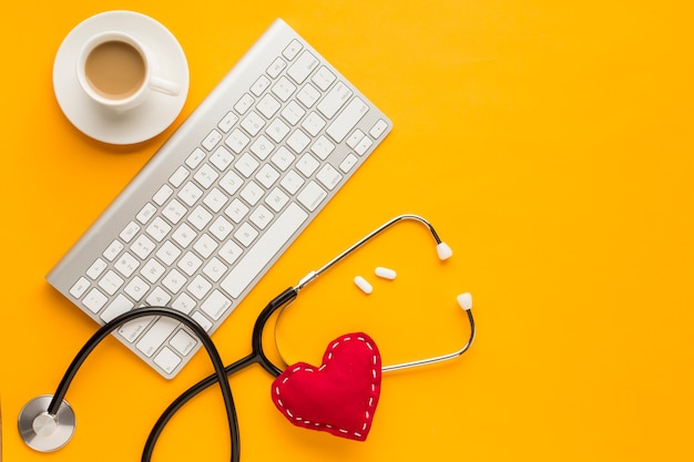 Free photo top view of wireless keyboard; tablets; coffee cup; stethoscope; stitched toy heart; above yellow backdrop