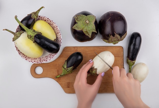 Top view  woman cuts black and white eggplant on a cutting board with a knife and in a plate  on a white background