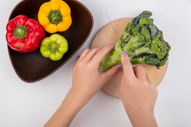 Top view  woman cuts broccoli on a cutting board with colored bell peppers in a bowl on a white background