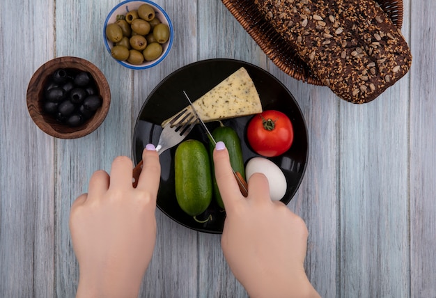 Top view  woman cuts cheese with cucumbers  tomato  egg on a plate with black and green olives on gray background