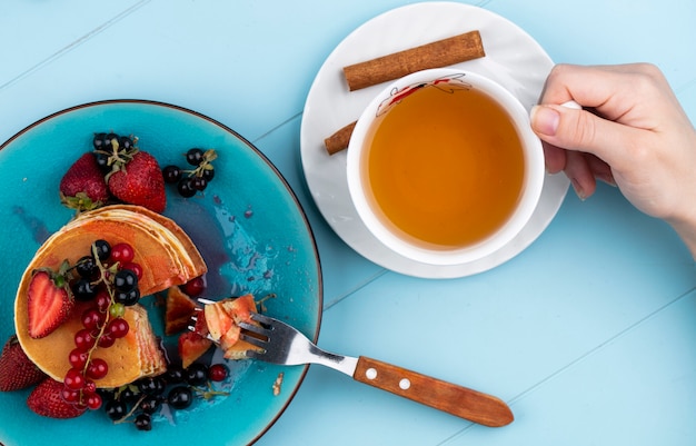 Top view of a woman drinks a cup of tea with pancakes with strawberries red and black currants on a blue surface