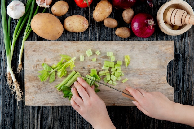 Top view of woman hand cutting celery on cutting board with vegetables and garlic crusher on wooden background