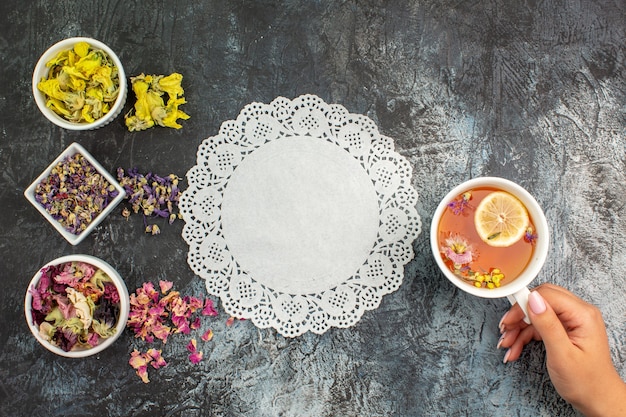 Top view of woman hand holding a cup of herbal tea and bowls of dry flowers with lace on grey ground