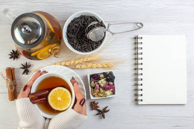 Top view of woman hand holding a cup with herbal tea with cinnamon lime lemon and necessary ingredients next to notebook on white background