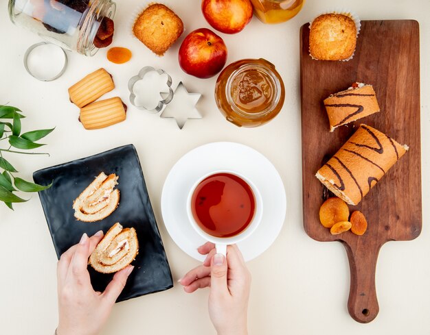 Top view of woman hands holding cup of tea and roll slice with jam, cookies, raisins and dried plums on white table