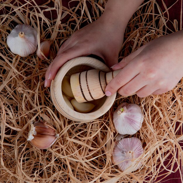 Top view of woman hands pressing garlic cloves in garlic crusher and garlic bulbs around on straw background