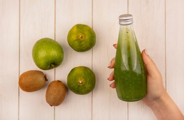 Top view woman holding juice bottle with apple kiwi and tangerines on white wall