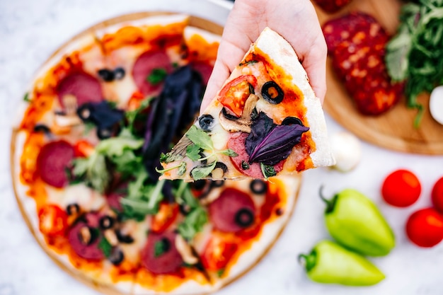 Top view of woman holding a slice of pepperoni pizza with olive tomato mushroom and herbs