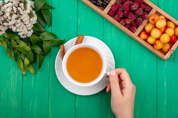 Top view woman holds cup of tea with cinnamon raspberries and white cherries on a green table