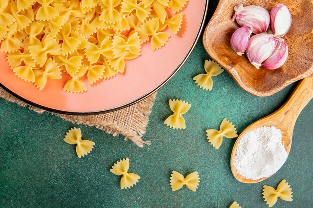 Top view wooden spoon with flour with raw pasta in a plate with garlic on a green table