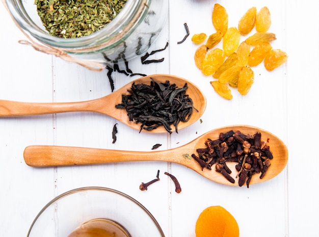 Free photo top view of wooden spoons with dry black tea leaves and clove spice and scattered dried raisins on white wood