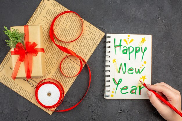 Top view xmas gift in brown paper branch fir ribbon on newspaper happy new year written on notepad pencil in female hand on dark surface