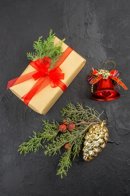 Top view xmas gift in brown paper tied with red ribbon fir branch xmas tree ornaments on dark surface