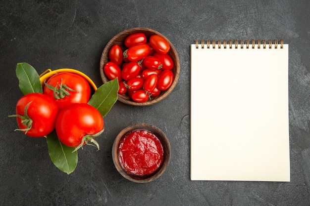 Free photo top view yellow bucket with tomatoes and bay leaves bowls with cherry tomatoes and ketchup and a notebook on dark ground