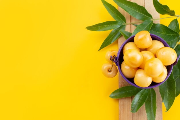 A top view yellow cherries mellow and sweet with green leaves on yellow desk, fruit color summer sweet cherry